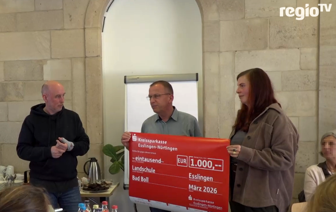 Drei Personen präsentieren einen großen roten Scheck bei einer Veranstaltung.